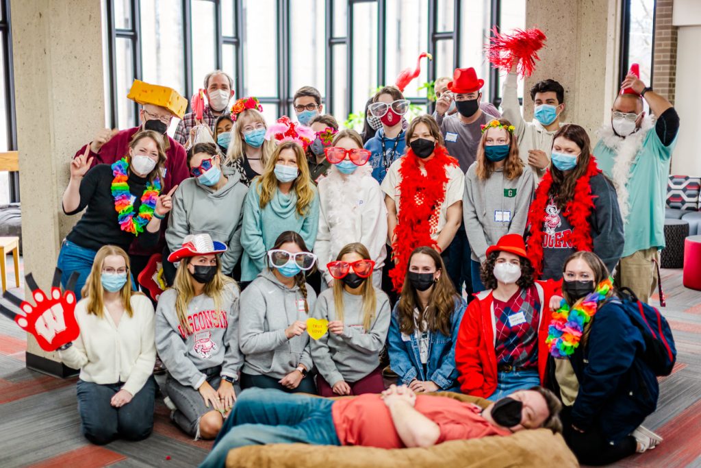 photo of MERIT staff members in the library atrium, wearing silly glasses, funny hats, and holding goofy props